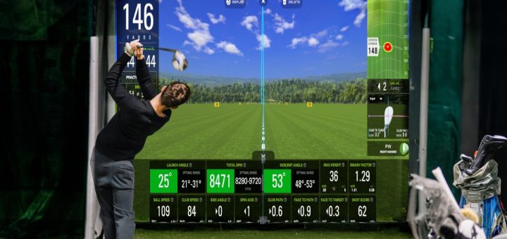 A golfer swinging a club indoors toward a digital golf screen with yardage metrics, green turf, and golf bags nearby.
