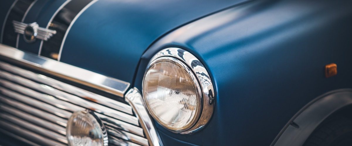 A blue vintage car fills the frame with a chrome headlight and grille as reflections sweep across the curved hood.