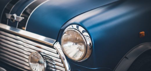 A blue vintage car fills the frame with a chrome headlight and grille as reflections sweep across the curved hood.