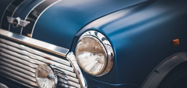 A blue vintage car fills the frame with a chrome headlight and grille as reflections sweep across the curved hood.