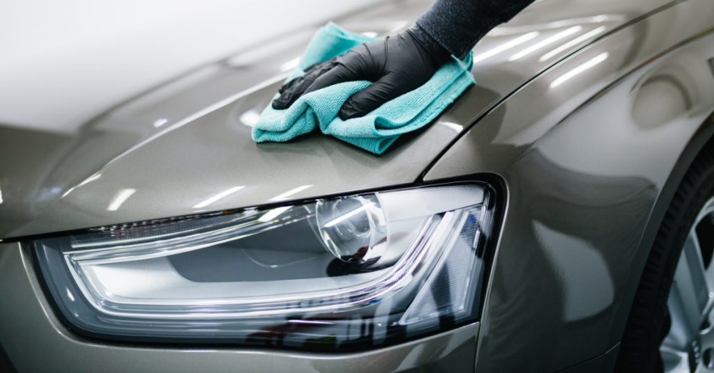 A person wiping the front of a car with a blue microfiber cloth near a headlight in a garage under indoor lighting.