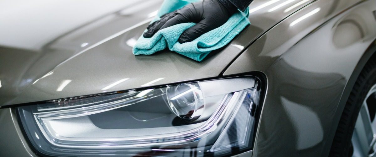 A person wiping the front of a car with a blue microfiber cloth near a headlight in a garage under indoor lighting.