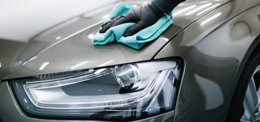 A person wiping the front of a car with a blue microfiber cloth near a headlight in a garage under indoor lighting.