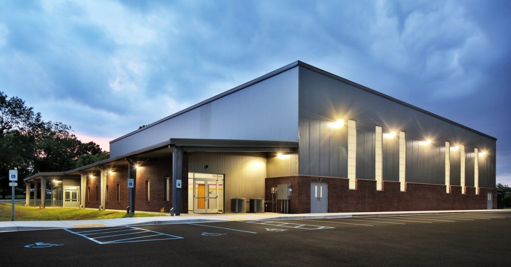 Modern commercial building with illuminated exterior lights, covered entrance, and empty parking lot beneath a cloudy sky.