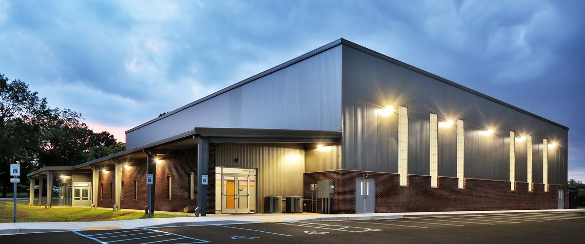 Modern commercial building with illuminated exterior lights, covered entrance, and empty parking lot beneath a cloudy sky.