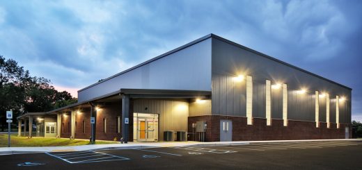 Modern commercial building with illuminated exterior lights, covered entrance, and empty parking lot beneath a cloudy sky.