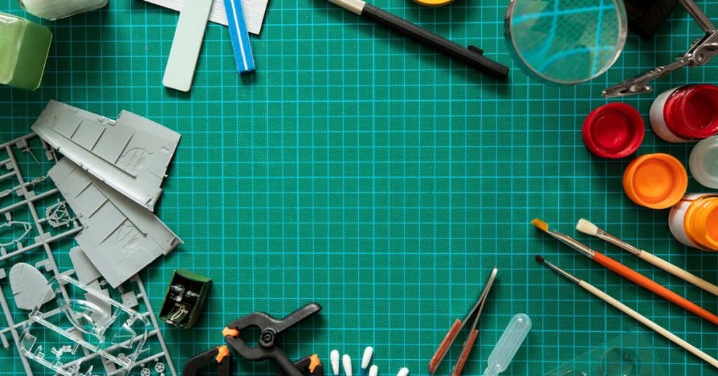 An overhead view of various hobby tools like paintbrushes, blades, and rulers on a green cutting mat.