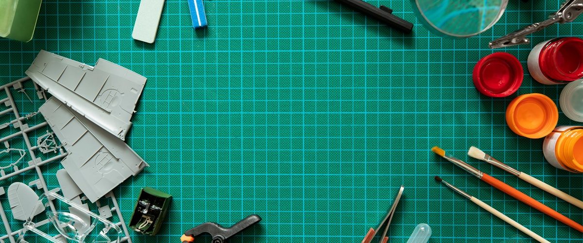 An overhead view of various hobby tools like paintbrushes, blades, and rulers on a green cutting mat.