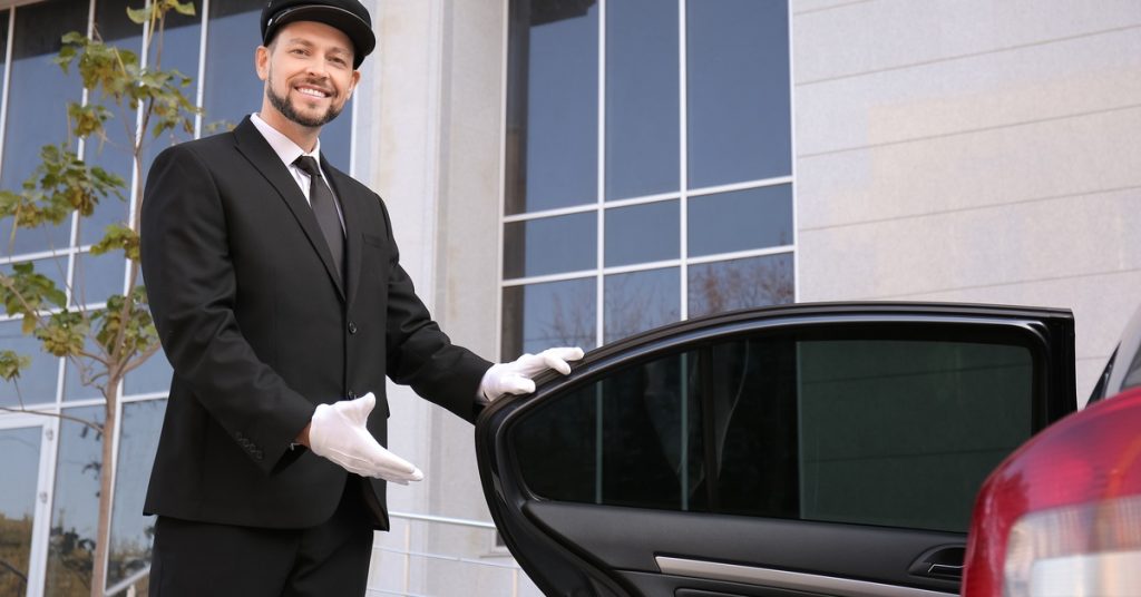 A man wearing a black suit and a black hat is smiling as he holds the back driver's side door open of a sedan.