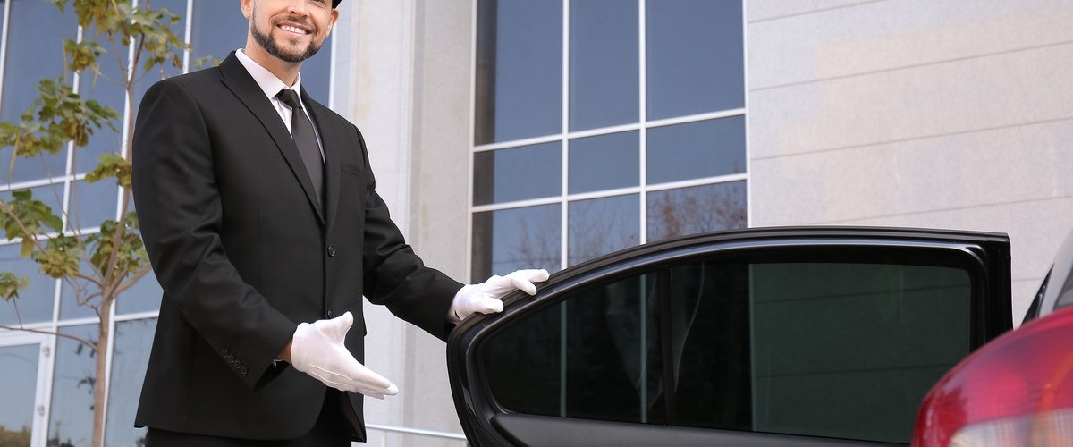 A man wearing a black suit and a black hat is smiling as he holds the back driver's side door open of a sedan.