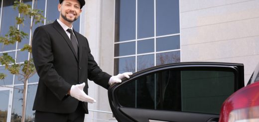 A man wearing a black suit and a black hat is smiling as he holds the back driver's side door open of a sedan.
