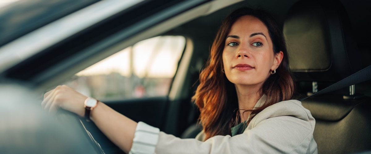 Woman seated behind the wheel of a car looks out the driver's side window with one hand resting on the steering wheel.