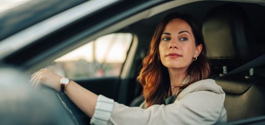 Woman seated behind the wheel of a car looks out the driver's side window with one hand resting on the steering wheel.