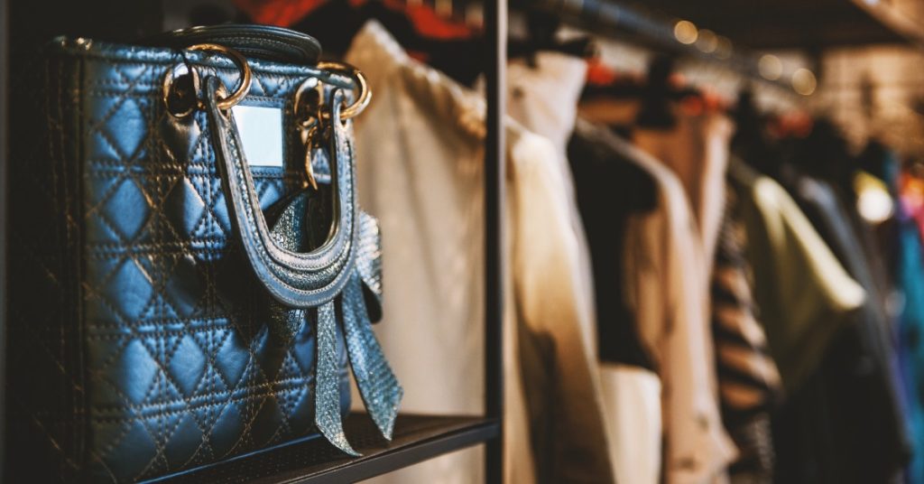 A close-up of a black leather bag with gold hardware placed on a shelf next to a clothing rack with multiple items.