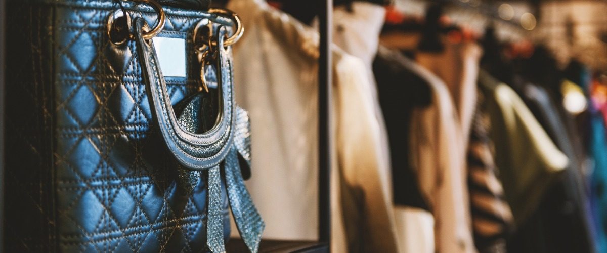 A close-up of a black leather bag with gold hardware placed on a shelf next to a clothing rack with multiple items.