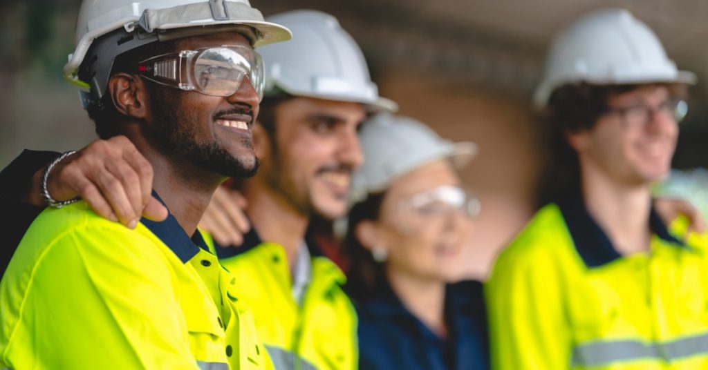 A group of construction workers in protective gear standing in a line with their arms on each other's shoulders.
