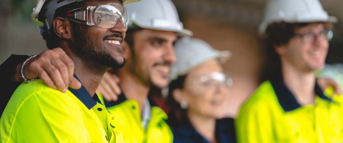 A group of construction workers in protective gear standing in a line with their arms on each other's shoulders.