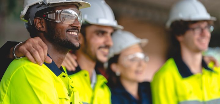 A group of construction workers in protective gear standing in a line with their arms on each other's shoulders.
