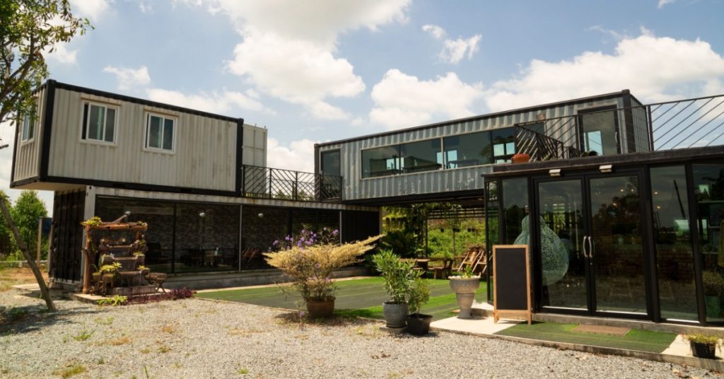 Modern building built from stacked shipping containers with large windows, glass entryway, patio plants, and open courtyard.