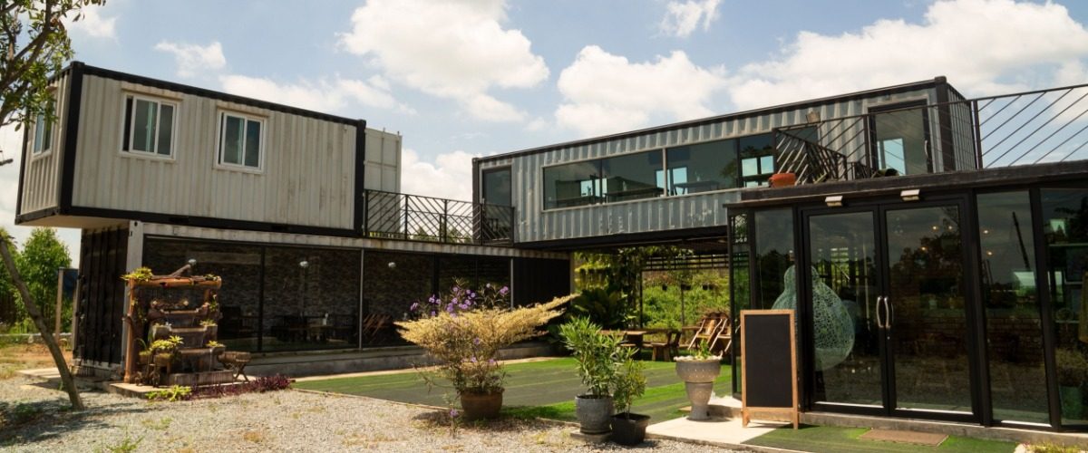 Modern building built from stacked shipping containers with large windows, glass entryway, patio plants, and open courtyard.