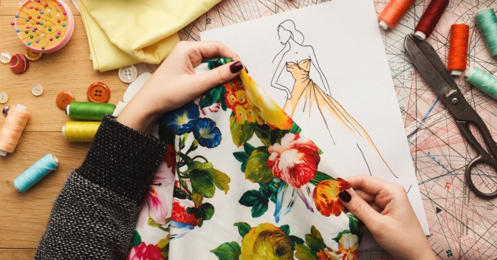 A close-up of two hands holding a floral piece of fabric over a sketch of a dress on a desk with scissors and thread.