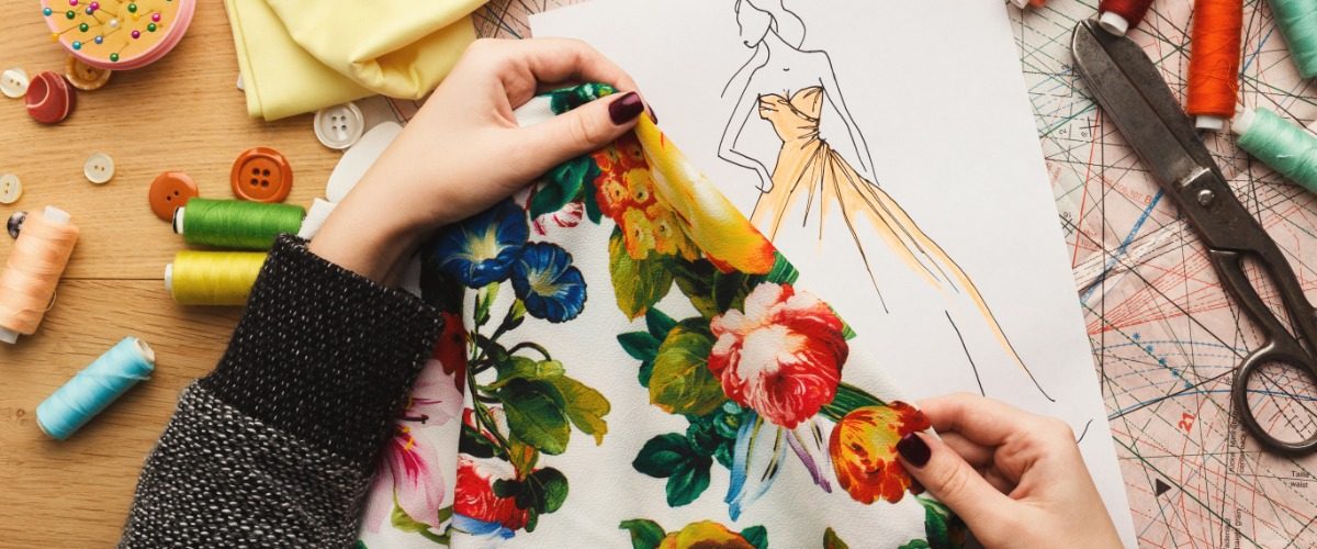 A close-up of two hands holding a floral piece of fabric over a sketch of a dress on a desk with scissors and thread.