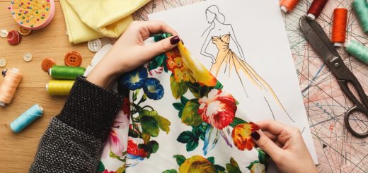 A close-up of two hands holding a floral piece of fabric over a sketch of a dress on a desk with scissors and thread.