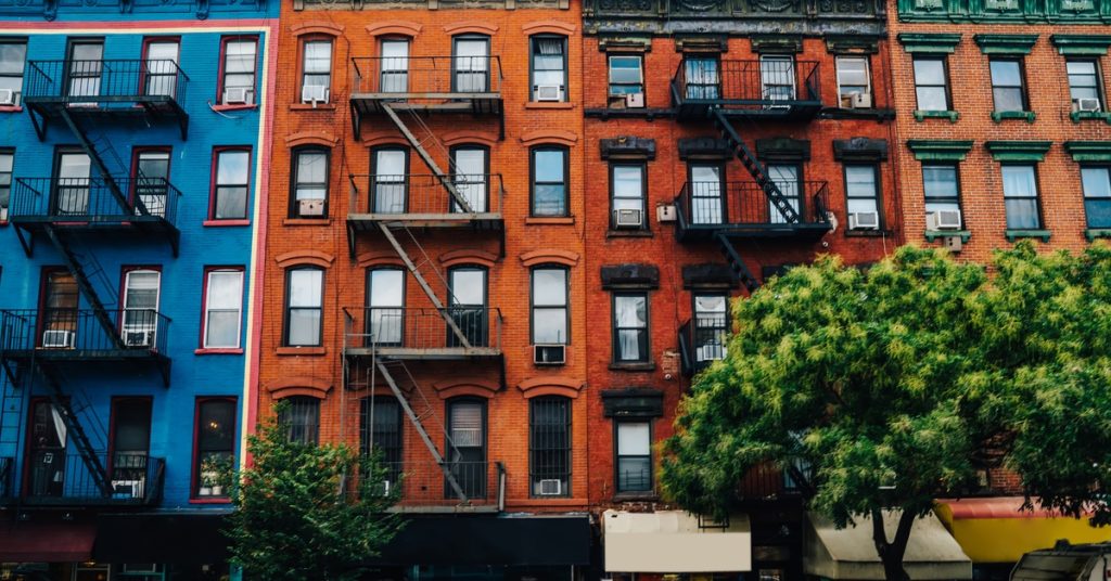 Row of blue, red, and green apartment buildings with fire escapes, storefronts, and leafy trees along the sidewalk.