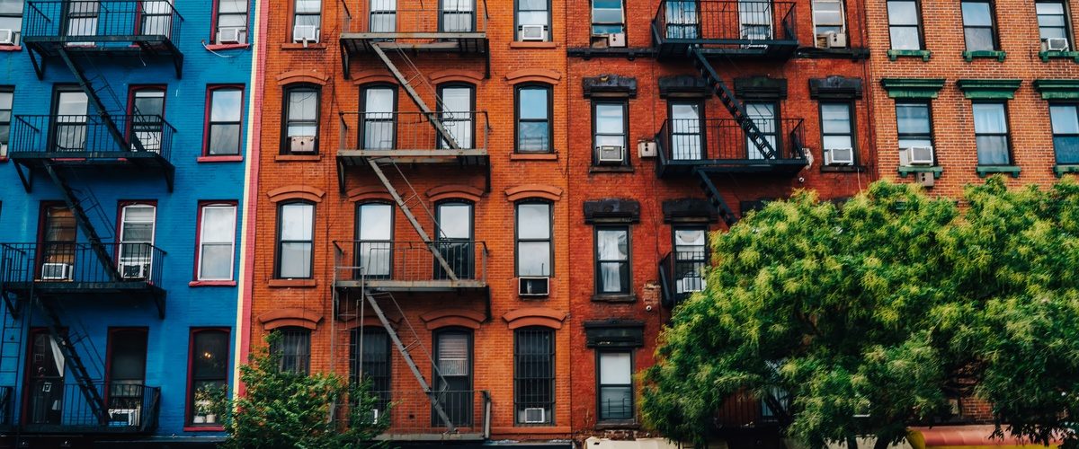 Row of blue, red, and green apartment buildings with fire escapes, storefronts, and leafy trees along the sidewalk.