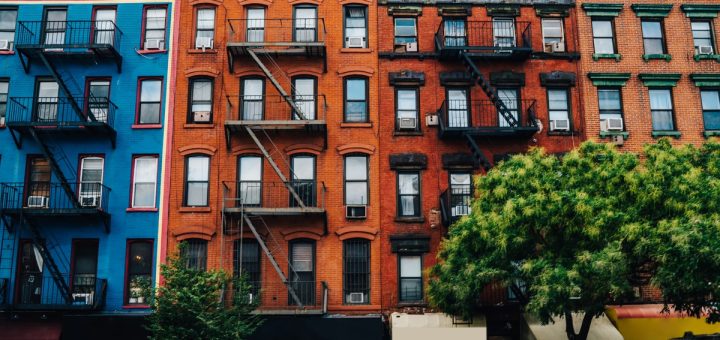 Row of blue, red, and green apartment buildings with fire escapes, storefronts, and leafy trees along the sidewalk.