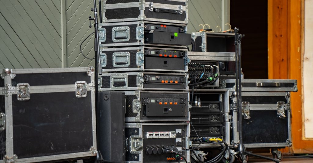A stack of black audio rack cases with power amplifiers and receivers on an outdoor stage with cables nearby.