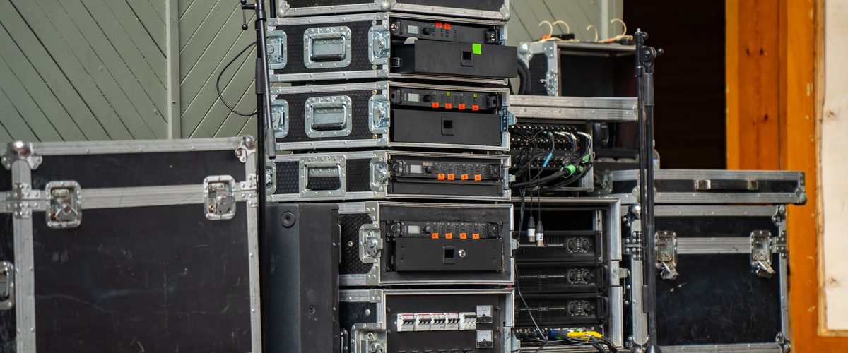 A stack of black audio rack cases with power amplifiers and receivers on an outdoor stage with cables nearby.