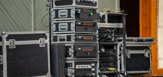 A stack of black audio rack cases with power amplifiers and receivers on an outdoor stage with cables nearby.