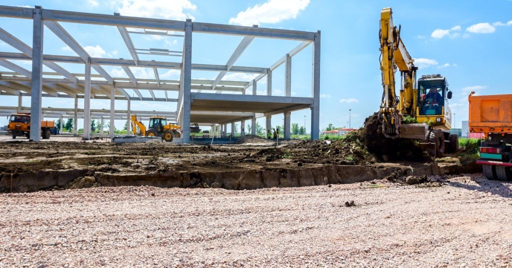 The frame of a building is built in the middle of an empty field. There is a large piece of machinery next to the frame.
