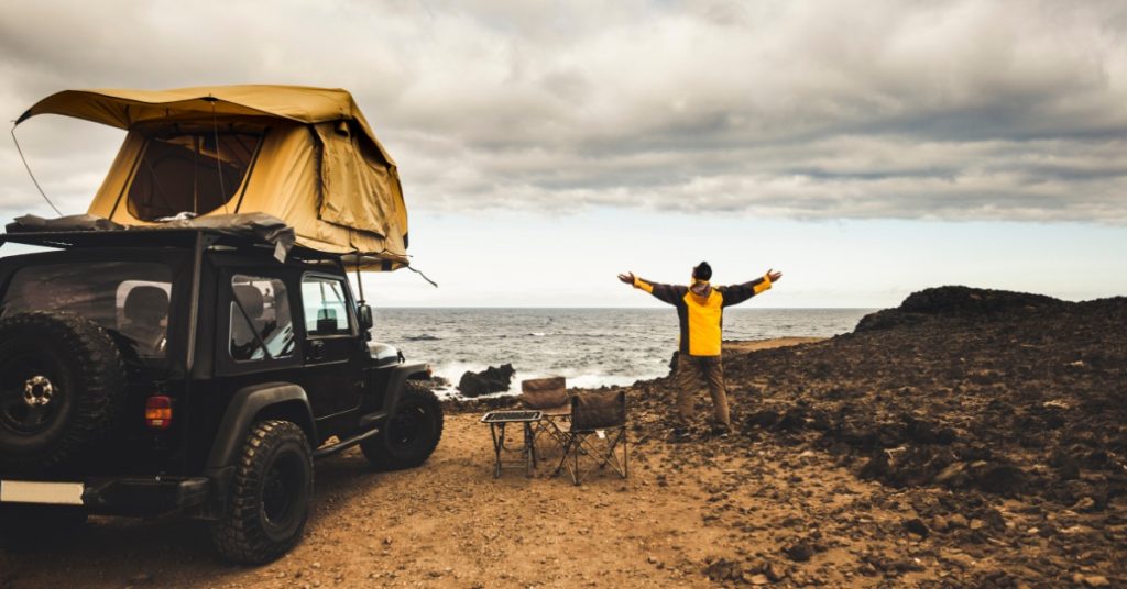 A man wearing a yellow jacket standing with both arms open next to a Jeep with a roof tent next to the ocean.