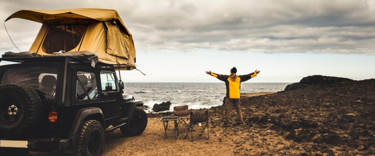A man wearing a yellow jacket standing with both arms open next to a Jeep with a roof tent next to the ocean.