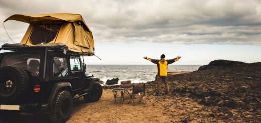 A man wearing a yellow jacket standing with both arms open next to a Jeep with a roof tent next to the ocean.