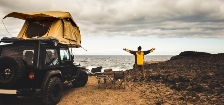 A man wearing a yellow jacket standing with both arms open next to a Jeep with a roof tent next to the ocean.