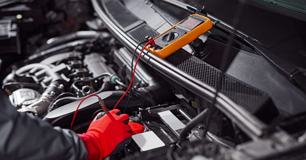 A mechanic wearing bright red gloves using a small orange tool to check the voltage of a car battery.