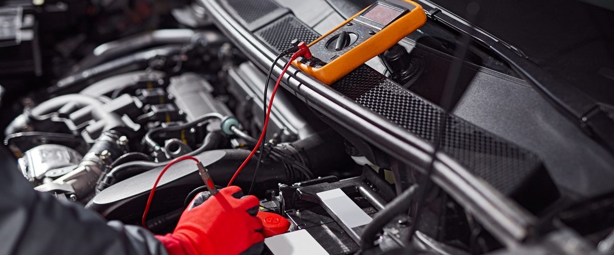 A mechanic wearing bright red gloves using a small orange tool to check the voltage of a car battery.
