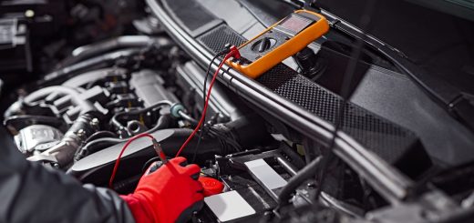 A mechanic wearing bright red gloves using a small orange tool to check the voltage of a car battery.