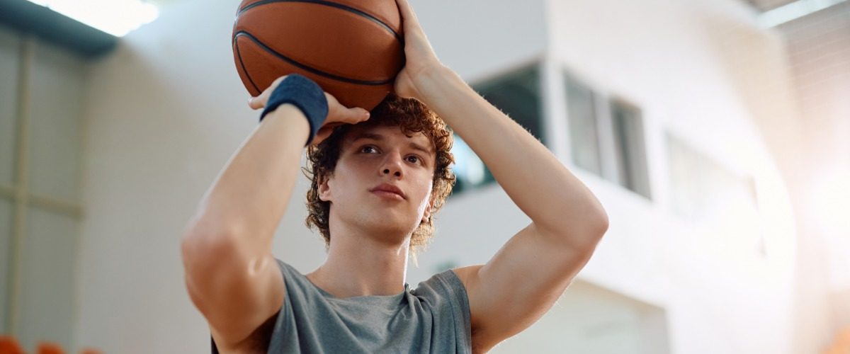 A close-up of a young man standing in a gym. He holds a basketball up near his forehead, ready to shoot a basket.