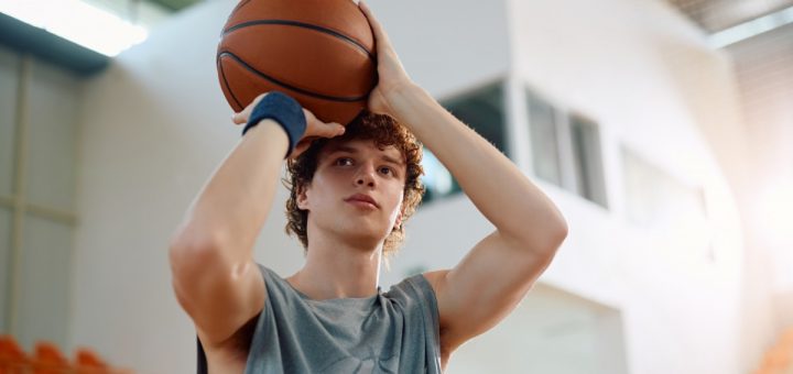 A close-up of a young man standing in a gym. He holds a basketball up near his forehead, ready to shoot a basket.