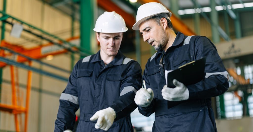 Two employees wearing white hard hats and white gloves inside a large, brightly lit industrial workspace.