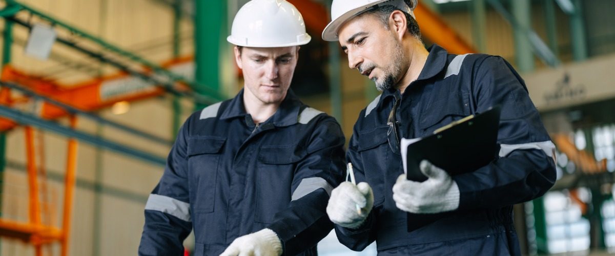Two employees wearing white hard hats and white gloves inside a large, brightly lit industrial workspace.