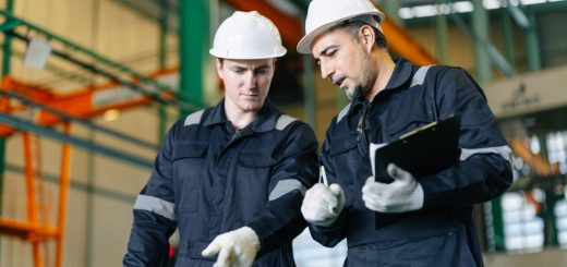 Two employees wearing white hard hats and white gloves inside a large, brightly lit industrial workspace.