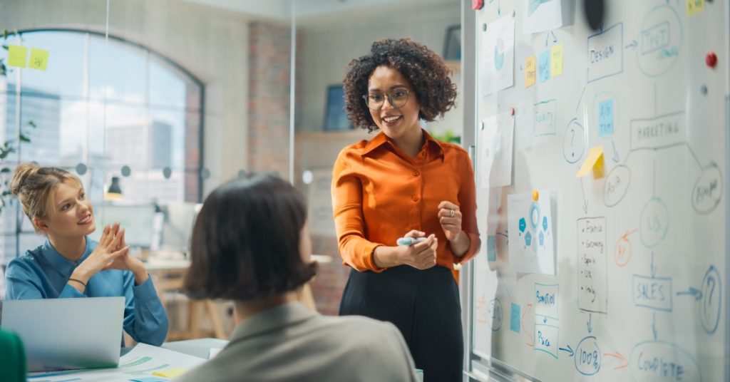Two employees sitting at a table watching another employee give a presentation in front of a dry erase board.