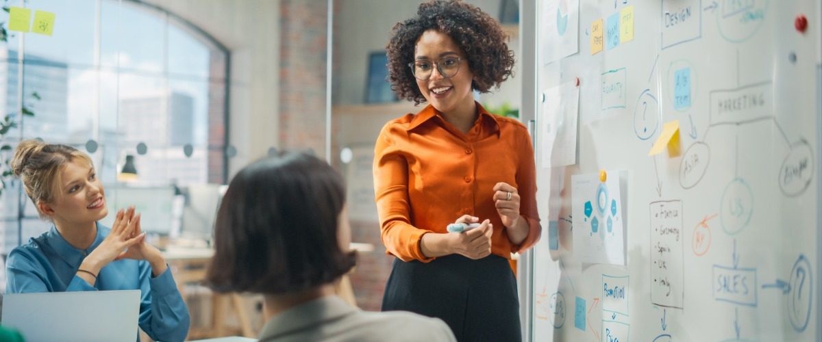 Two employees sitting at a table watching another employee give a presentation in front of a dry erase board.