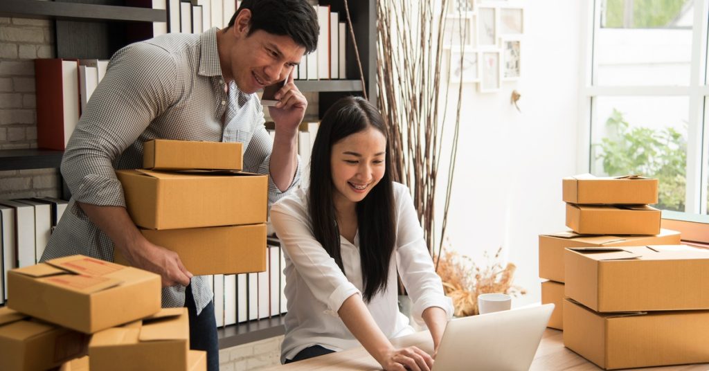 A man holding packages while on the phone as a woman works on a laptop surrounded by shipping boxes.
