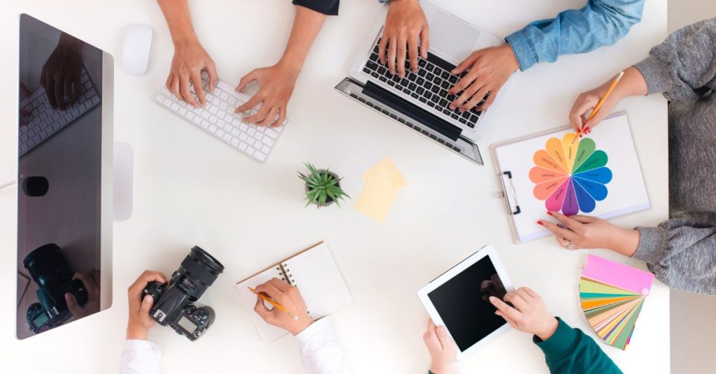A top view of four people sitting around a table with laptops, papers, drinks, and notebooks in a bright office.